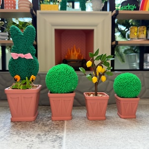 May include: A collection of miniature potted plants. Includes a green bunny-shaped topiary with a pink bow, two round green topiary plants, and a small lemon tree with yellow fruit. All plants are in pink square pots.