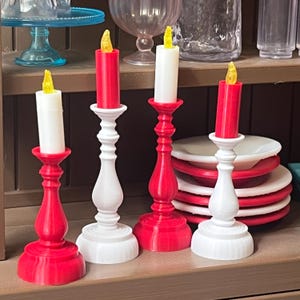 May include: Four decorative candle holders with red and white bases and faux flickering yellow flames. The candle holders are arranged on a wooden shelf, with a stack of red-rimmed plates in the background.