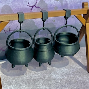 May include: Three black cauldron-shaped hanging pots with hooks. The pots are different sizes and are hanging from a wooden bar.