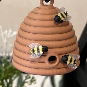 Op de afbeelding: Een bruin, bijenkorfvormig ornament met drie decoratieve bijen. De bijen hebben zwarte lichamen, gele strepen en doorzichtige vleugels. De bijenkorf heeft een lus aan de bovenkant om op te hangen.