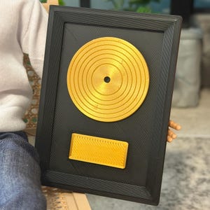 May include: A rectangular black plaque featuring a centered circular gold record and a small rectangular gold nameplate at the bottom. The plaque has a textured, lined surface finish throughout, with a raised border framing the entire piece.