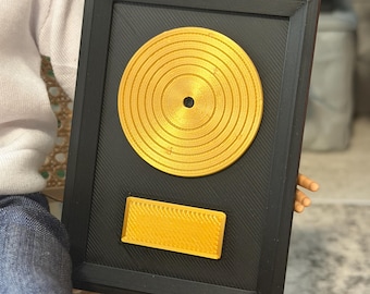 May include: A rectangular black plaque featuring a centered circular gold record and a small rectangular gold nameplate at the bottom. The plaque has a textured, lined surface finish throughout, with a raised border framing the entire piece.
