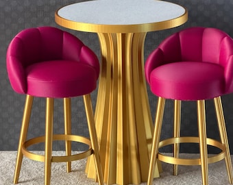 May include: A round table with a white speckled top and a gold fluted pedestal base, flanked by two bar stools featuring bright magenta upholstered seats with curved backrests and gold metal legs with circular footrests.