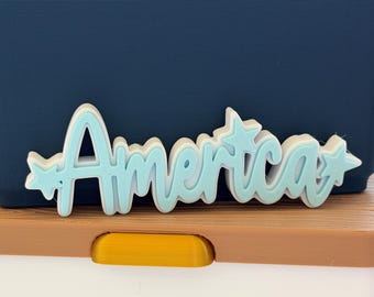 May include: A light blue decorative charm featuring the word America in a cursive script font. The design includes a small five-pointed star on the far left and a larger five-pointed star on the far right, with a white layered base underneath.