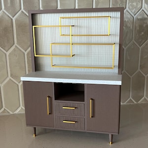 Op de afbeelding: Een bruin houten kastje met een wit aanrechtblad en gouden metalen planken. De kast heeft twee deuren met verticale groeven en twee laden met gouden metalen handvatten. De planken zijn in een geometrisch patroon gerangschikt.