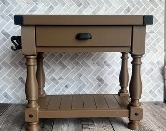 May include: A brown wooden kitchen island with a drawer and a lower shelf. The island has turned legs and black metal corner brackets and a handle. The island is 30 inches long, 18 inches wide, and 36 inches high.