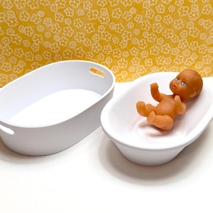 Peut inclure: Un petit poupon brun aux yeux bleus est allongé dans un bain blanc ovale. Un autre récipient blanc ovale est à côté du bain. Le fond est jaune avec un motif floral.