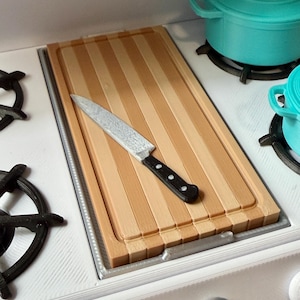 May include: A miniature kitchen scene featuring a wooden cutting board with a stainless steel knife and black handle. A teal pot sits on a white stove with black burners. The cutting board is made of light brown wood.