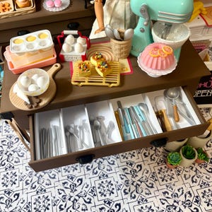 May include: A miniature kitchen scene with a brown cabinet, open drawer with utensils, and various baking accessories. Includes a pink cake, a teal mixer, and a cutting board with eggs. The scene is set on a patterned surface.