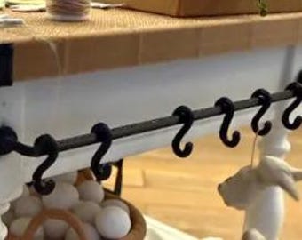 May include: A black metal rod mounted horizontally to a white wooden structure, featuring six black S-shaped hooks hanging from the bar. The rod is secured at both ends with circular black brackets, providing a functional storage solution.