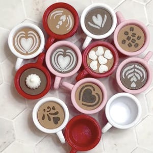 May include: A collection of miniature coffee cups in various colors, including red, pink, and white. Each cup features a different latte art design, such as hearts, leaves, and coffee beans. The cups are arranged in a circular pattern.