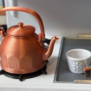 May include: A copper-colored teapot with a black base and a white teacup with a flower design. The teacup has a tea bag attached. The teapot is on a white stove top.