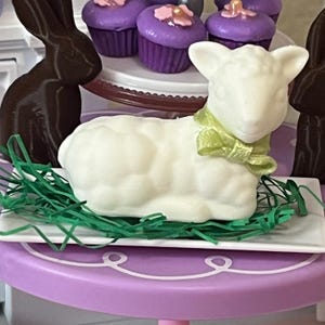 May include: A white chocolate lamb figurine with a green bow, lying on a bed of green grass, on a white plate.