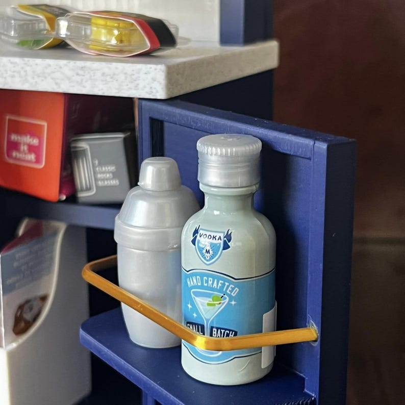 Op de afbeelding: Een miniatuur barset met een zilveren cocktailshaker en een klein flesje wodka. De fles heeft een blauw etiket met de tekst "VOOKA HAND CRAFTED SMALL BATCH".