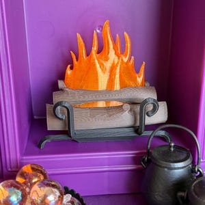 May include: A miniature fireplace scene featuring a translucent orange flame insert resting on brown logs. The logs are held by a black metal-style grate with scrolled ends. A small black cauldron sits to the right, and iridescent spheres are in the foreground.
