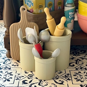 May include: Four beige ceramic containers with a variety of kitchen utensils, including wooden rolling pins, spoons, and a pastry brush. The containers are on a black and white tiled surface.