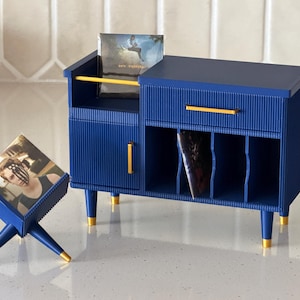 May include: A miniature navy blue storage cabinet with gold accents, featuring a fluted texture, a small drawer, and open shelving for vinyl records. Beside it sits a matching X-shaped stand holding a square album cover featuring a person's portrait.