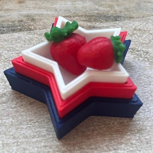 May include: A patriotic star-shaped dish in red, white, and blue, layered to create a tiered effect. Two red strawberries with green stems rest inside the white layer, adding a touch of freshness to the design.