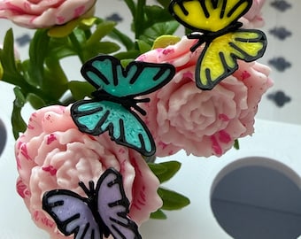 Peut inclure: Gros plan sur une composition florale avec des pivoines roses et trois décorations de papillons colorés. Les papillons sont turquoise, jaunes et violets avec des contours noirs. L'arrangement est sur fond blanc.