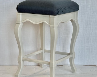 May include: A small stool featuring a cream-colored base with four curved legs and a square, dark charcoal-colored seat. The frame has a scalloped apron design connecting the legs, and the seat surface displays a fine, horizontal ribbed texture.