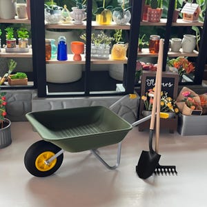 May include: A miniature garden scene featuring a green wheelbarrow with a yellow wheel, a black shovel and rake, and a greenhouse filled with plants and flowers. The scene is set on a white surface.