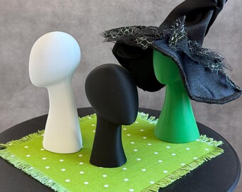 May include: Three abstract head-shaped display stands in white, black, and green. The green head wears a black pointed hat with a black lace trim. The heads are arranged on a square lime green fabric mat featuring a white polka dot pattern.