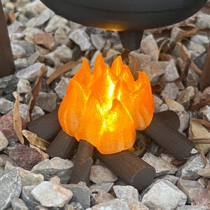 May include: A miniature campfire with bright orange flames and dark brown logs. The flames are lit, and the logs are arranged in a classic campfire style. The scene is set on a bed of small rocks, suggesting an outdoor setting.