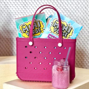 May include: A magenta rubber tote bag with perforated holes and two handles, containing multiple MGA's Miniverse boxes with blue and yellow graphic text. A small clear glass jar with a pink smoothie and a straw sits in front of the bag.