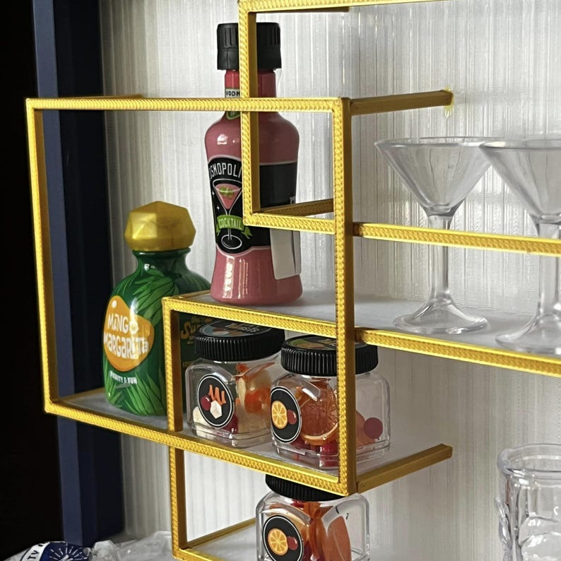 Op de afbeelding: Een gouden metalen rek met meerdere planken. De planken zijn gevuld met verschillende items, waaronder twee flessen drank, twee martini glazen en verschillende kleine potten gevuld met kleurrijke snoepjes. Het rek staat tegen een witte achtergrond.