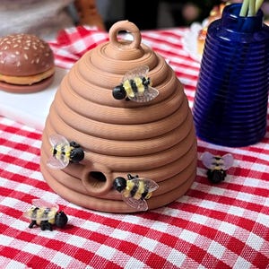 Puede incluir: Un objeto decorativo en forma de colmena marrón con un lazo en la parte superior y varias figuras de abejas. Las abejas tienen rayas amarillas y negras y alas transparentes. El artículo está sobre un mantel a cuadros rojos y blancos.