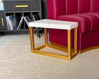 May include: A rectangular side table featuring a white speckled top and a gold-colored metal frame. The table has a clean, geometric design with four vertical legs connected by a base frame, positioned in front of a deep magenta sofa and a dark cabinet.