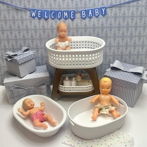 Puede incluir: Una escena con temática de bebé con muñecas en miniatura, una cuna y cajas de regalo. La escena incluye una pancarta que dice "WELCOME BABY". Las muñecas están en una bañera y una cuna. Las cajas de regalo son grises con lunares blancos y lazos.
