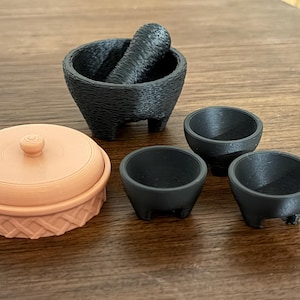 May include: A collection of small kitchen tools, including a black mortar and pestle, three small black bowls, and a peach-colored round container with a lid. The items are arranged on a wooden surface.