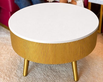 May include: A round coffee table featuring a smooth white top and a cylindrical base with a vertical ribbed texture in a metallic gold finish. The table is supported by three short, tapered legs in a matching gold color.