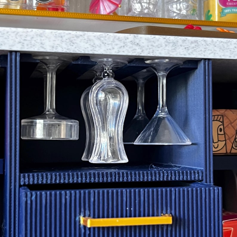 Op de afbeelding: Een set van vier heldere glazen wijnglazen die aan een donkerblauwe rek hangen. De glazen zijn van verschillende vormen en maten. De rek is gemonteerd op een kast met een gele handgreep.