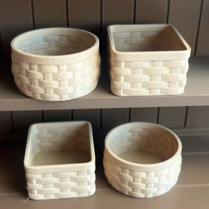 May include: Four off-white woven baskets of varying shapes and sizes. Two are round, and two are square. The baskets are arranged on a brown shelf. The woven pattern is visible on the sides of each basket.