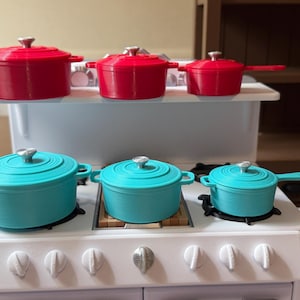 May include: A set of miniature red and teal-colored cast iron pots and pans. The set includes three red pots with lids and handles, and three teal pots with lids and handles. The pots are arranged on a white toy stove.