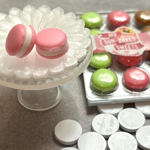May include: Two pink macarons with white filling sit on a small, clear glass cake stand. Several green and red candies are packaged in a clear plastic container. The packaging has the text "Les Sun-Baked Sweets".