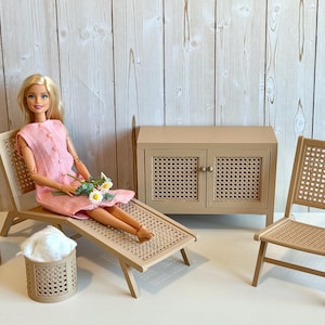 May include: A doll in a pink dress is seated on a miniature wooden chaise lounge, holding flowers. A matching side table with a drink and a small storage cabinet are nearby. A small chair and a basket are also present. The furniture has a woven design.
