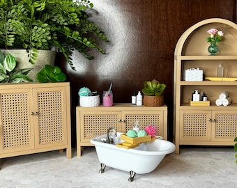 May include: A miniature bathroom scene with a white clawfoot bathtub, a light brown cabinet with a woven door, and a matching arched shelf. The scene includes decorative plants, bath products, and a drink in a glass.
