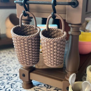 May include: Two small, woven brown baskets with rope handles hang from a brown wooden structure. The baskets have a textured, basket-weave design. The structure has a dark metal bar and is part of a larger, brown wooden piece.
