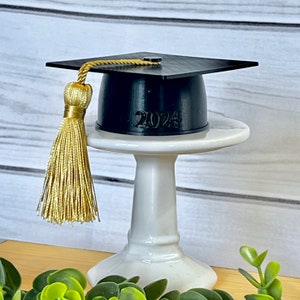 May include: Black graduation cap with a gold tassel and the year 2024 printed on the side. The cap is sitting on a white cake stand.