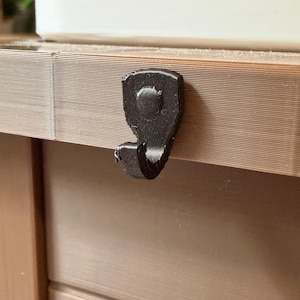 May include: A close-up of a dark gray hook attached to a brown wooden surface. The hook is made of a textured material and is designed to hold an item. The wooden surface has a grooved texture and appears to be part of a cabinet or furniture piece.