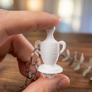 May include: A white, miniature vase-shaped keychain. The keychain features a small, detailed replica of a vase with handles and a textured surface. The keychain is attached to a silver keyring.