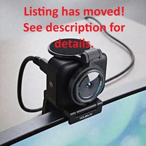 May include: A black YoloLiv webcam mounted on the top edge of a monitor. The device features a circular lens, a power button icon on the side, and a black cable connected to the back. Red text overlay reads: Listing has moved! See description for details.