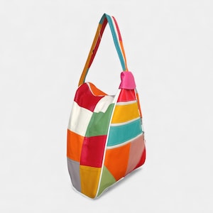 Könnte beinhalten: Bunter Patchwork-Schulterbeutel mit einem einzelnen Riemen. Die Tasche hat eine Vielzahl von Farben, darunter Rot, Weiß, Grün, Orange, Gelb und Türkis. Der Riemen ist mehrfarbig mit einem rosa Akzent oben.
