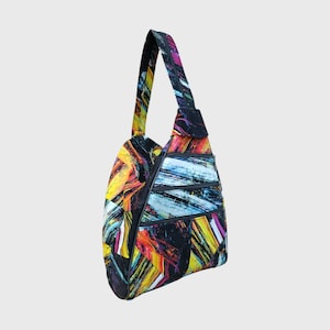 Könnte beinhalten: Eine farbenfrohe Schultertasche mit einem lebendigen abstrakten Design. Die Tasche hat eine schwarze Basis mit kräftigen gelben, orangefarbenen, blauen und weißen Strichen. Sie hat einen einzelnen Schulterriemen und mehrere Reißverschlussfächer.