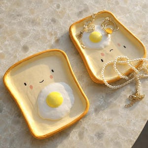 May include: Two rectangular ceramic trays shaped like slices of toast with rounded corners. Each tray features a hand-painted smiling face with pink cheeks and a raised, three-dimensional fried egg in the center. One tray holds a gold ring and necklace.