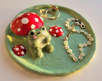 Puede incluir: Un plato decorativo redondo de cerámica verde claro con una rana verde esculpida, dos setas rojas con puntos blancos y un sombrero de seta rojo más pequeño. Un anillo dorado y una pulsera de cuentas con flores blancas descansan sobre el plato.
