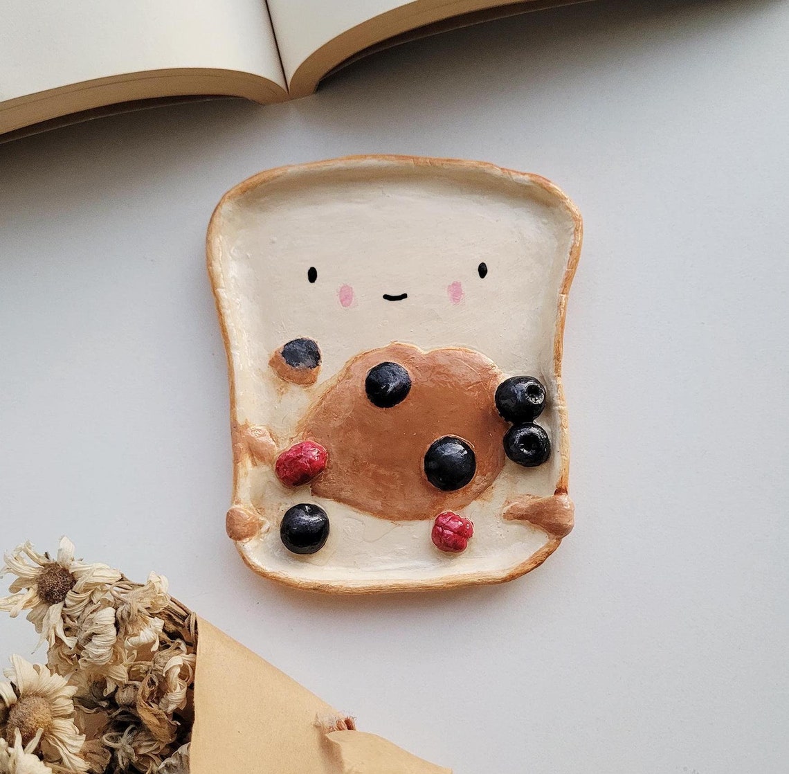 May include: A charming handmade clay trinket dish shaped like a slice of toast with a happy face.  The toast is off-white, and features a brown peanut butter spread with blueberries and raspberries. The blueberries are dark blue, and the raspberries are a deep red. This cute and whimsical dish is perfect for rings, earrings, or other small items.
